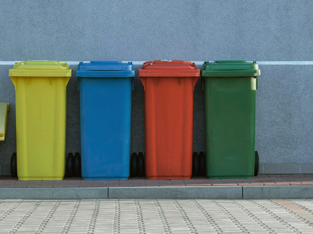 Vier Mülltonnen in gelb, blau, rot und grün auf dem Bürgersteig vor einer Wand