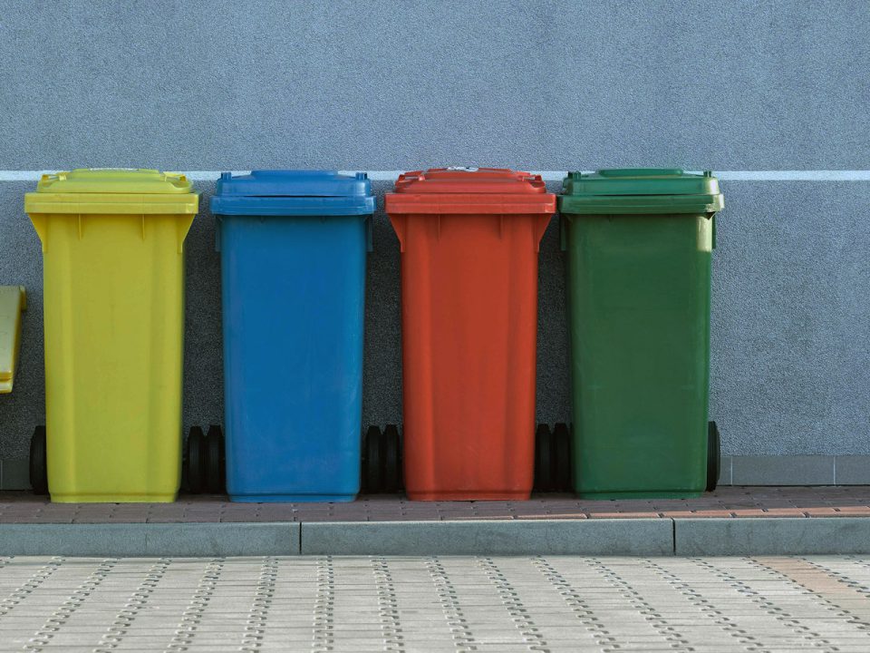 Vier Mülltonnen in gelb, blau, rot und grün auf dem Bürgersteig vor einer Wand